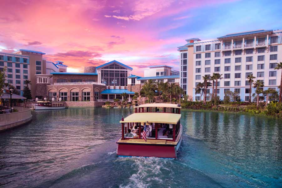 Water taxi traveling along a lagoon in front of Loews Sapphire Falls Resort, with the hotel buildings and tropical landscaping in the background.