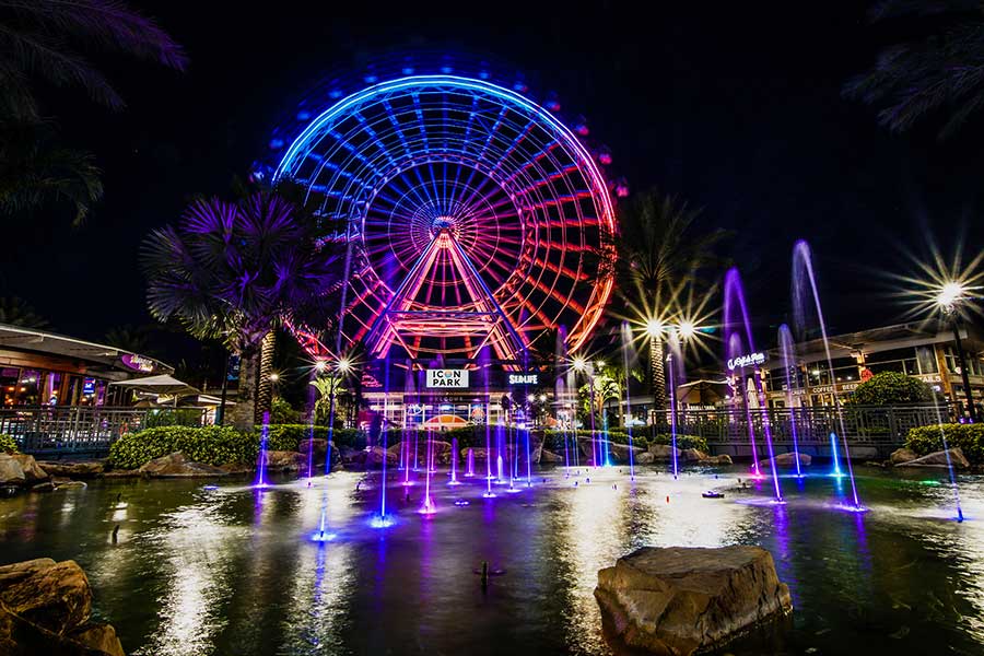Illuminated Ferris wheel at ICON Park reflected in the water at night, surrounded by buildings and colorful lights.